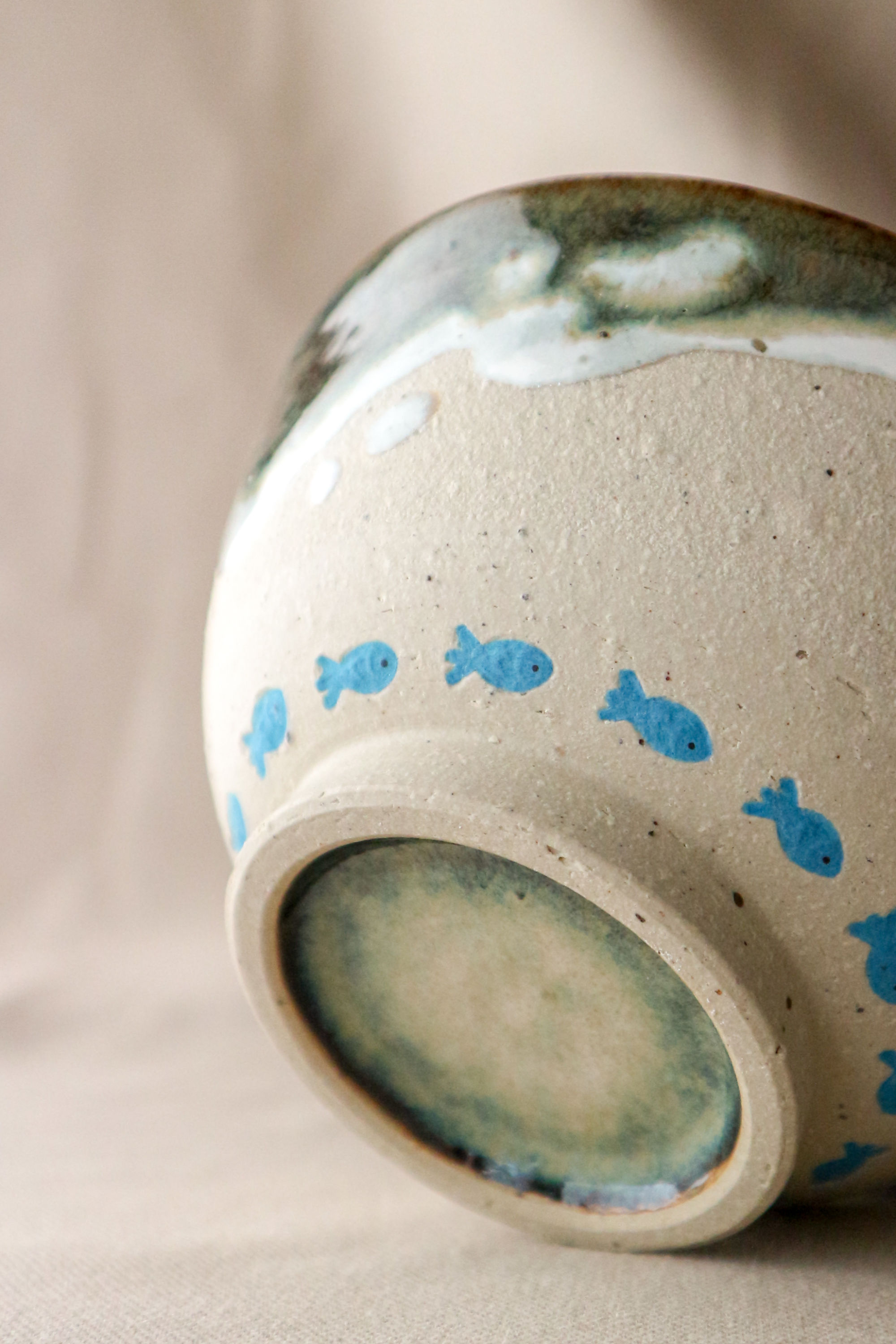 Hand-thrown stoneware ceramic bowl by Eden Hoang. Blue fish are embossed in a row along the base of the bowl, just above the foot. The bottom of the bowl is left unglazed to reveal the raw clay texture, with the top of the bowl glazed in a mixture of white & deep blue to represent the movement of waves.