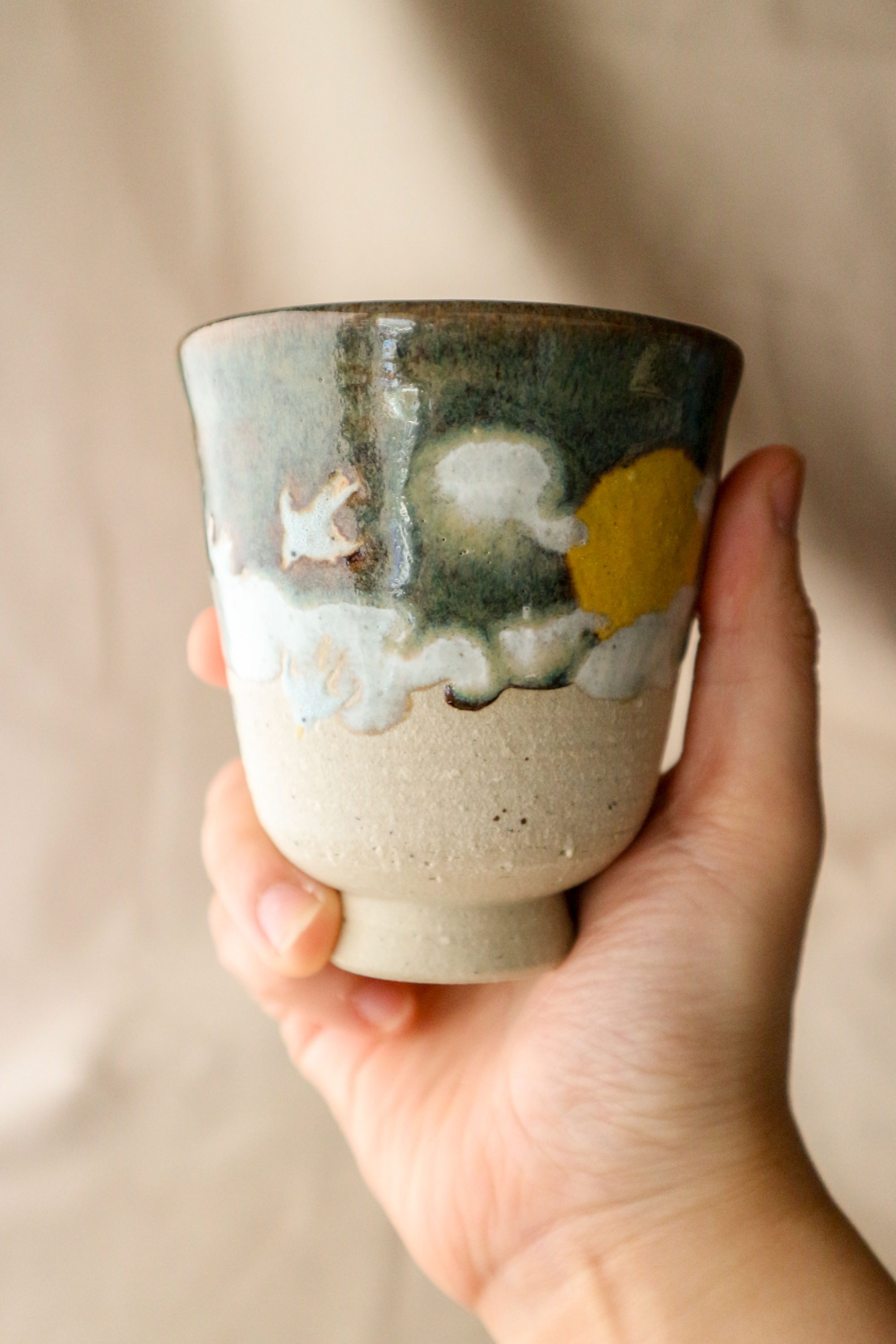 Hand-thrown stoneware ceramic cup by Eden Hoang. The base of the cup is left unglazed, revealing the raw clay texture. The top of the cup is decorated in a blue and white glaze, representing clouds in the night sky. There are also birds in flight, and a yellow moon sitting amongst the cloudy sky.
