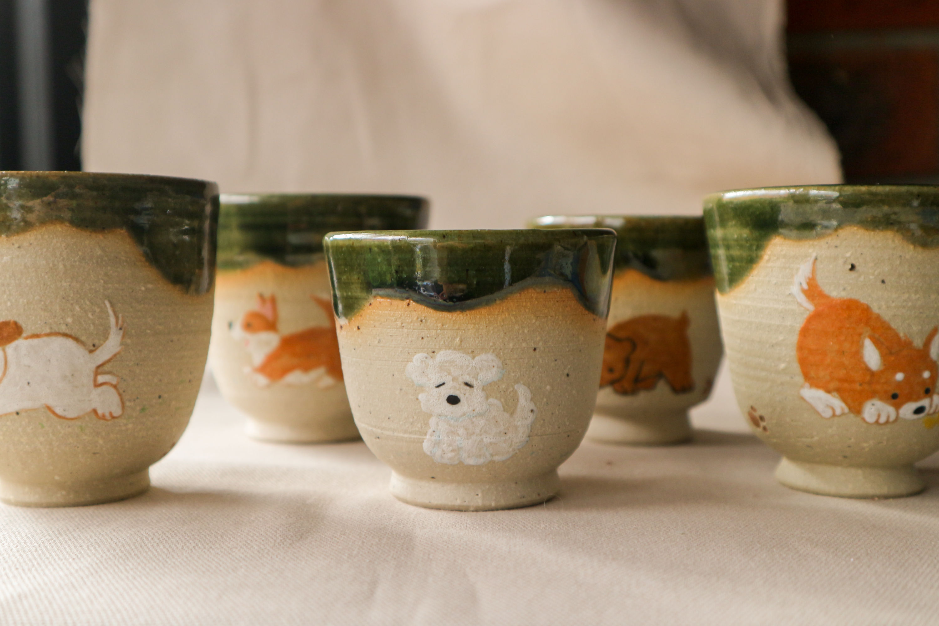 Hand-thrown stoneware ceramic cups by Eden Hoang. There are five cups visible. Four of them have various dog designs including a terrier, a maltese, a corgi and a shiba inu. One of them has a bear design. All of the cups have a deep forest green glaze dripping down the top rim of the cup.