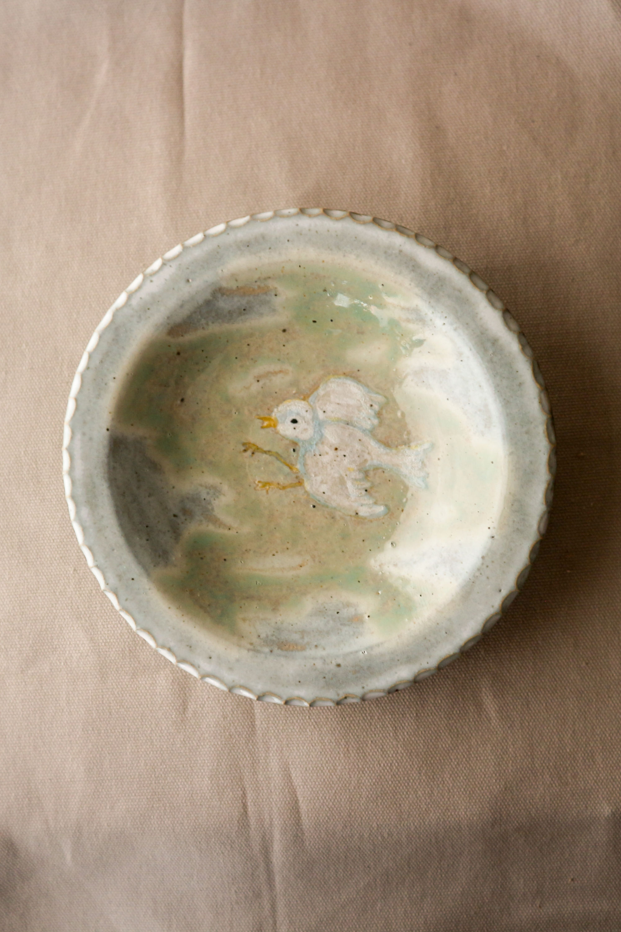 Hand-thrown stoneware ceramic plate by Eden Hoang. The rim of the plate is flat, with a textured patterned edge. At the centre of the plate, there is a white bird in flight amongst a pastel green cloudy sky. 