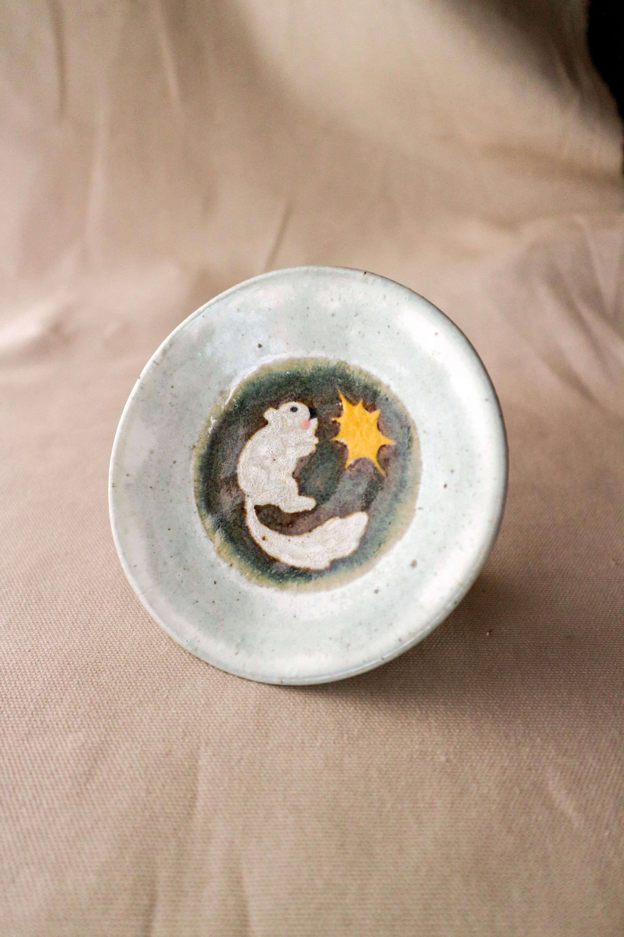 Hand-thrown stoneware ceramic plate by Eden Hoang.  The edge of the plate is glazed white. At the centre of the plate it is glazed in a deep blue, with a motif of a white fluffy rodent amazed at a shining star.