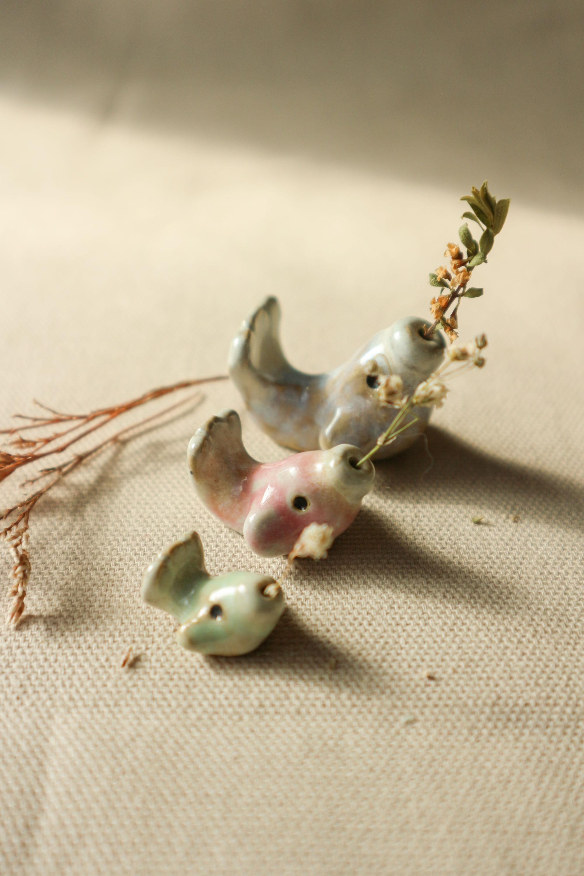 Hand-thrown stoneware ceramic fish figurines by Eden Hoang. Three fish figurines sit in a row, from smallest to largest. Smallest is light green, medium is soft pink, and largest is fluorescent blue. Each fish has a small flowered stem nestled in the mouth (there is a hole in the mouth for small stemmed flowers to be inserted).
