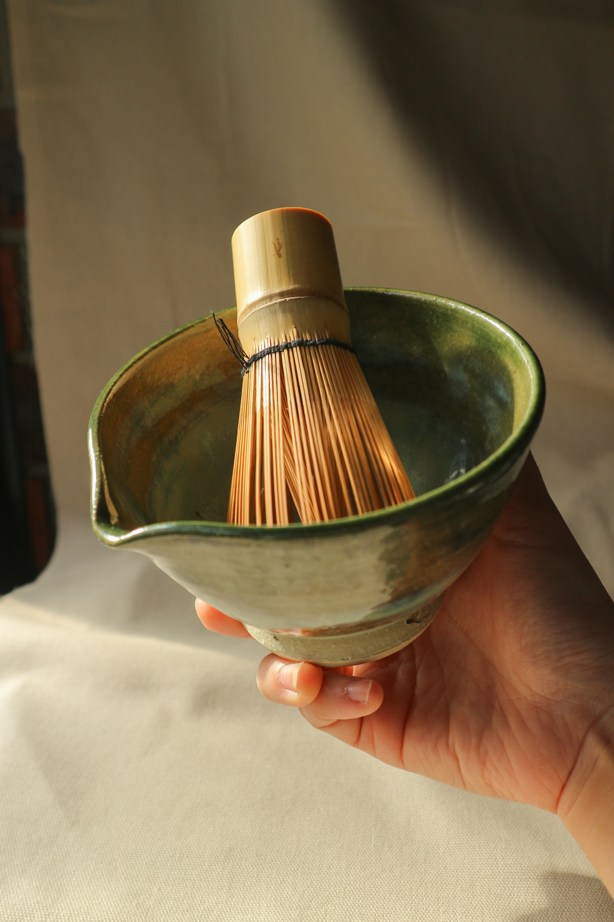 Hand-thrown stoneware ceramic matcha bowl by Eden Hoang. Held in the sunlight, the bowl is glazed in a deep forest green, with a matcha whisk resting inside the bowl.